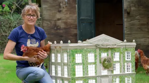 British Hen Welfare Trust (BHWT) A woman holding a hen next to a model house with a banner on the front saying 'The Millionth Hen'