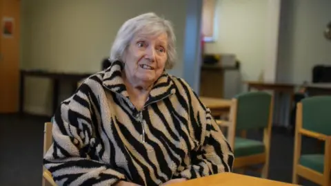 Una mujer, vestida con un polar beige con rayas negras y cremallera, está sentada a una mesa con los brazos extendidos. Al fondo, tras ella, se ven sillas verdes y algunos escritorios.