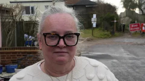 John Fairhall/BBC A head and shoulders image of Julia Durrant, who is standing in the road outside her home, although her home is not in the image. She is wearing a white jumper and glasses. She is looking directly into the camera.