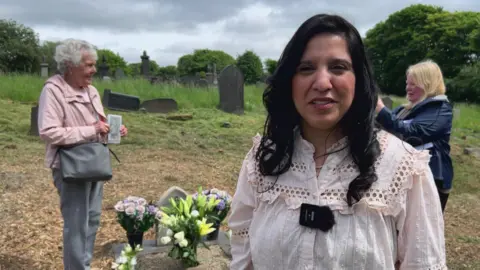 Nathan Turvey/BBC A picture of Yasmin Hanif, from the charity Barnardo's in Bradford, she has black hair and is wearing a white top. The grave is behind her with Pat holding a picture of her uncle by it in the background.