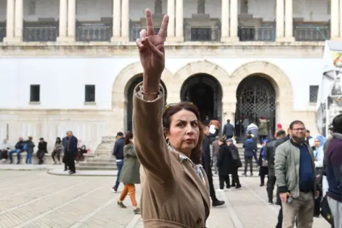 AFP A woman in a camel coat walks through a courtyard towards a municipal building. Her hand is raised in a 'V' sign.