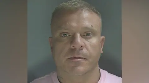 Custody photo of a man with short light hair and a pink t-shirt