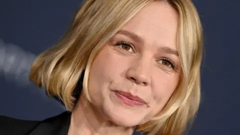 Carey Mulligan smiling at the camera, she has short blonde hair, brown eyes and a black jacket, with a navy blue background. 