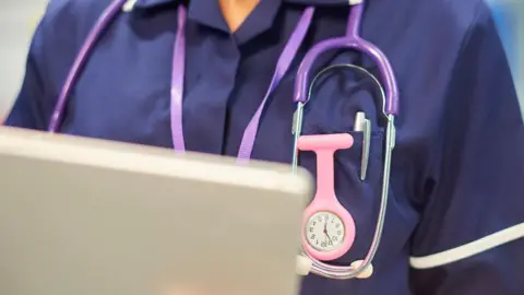 Getty Images A close-up of a nurse in purple scrubs wearing a stethoscope, using an iPad. Her chest area is visible, but not her face.