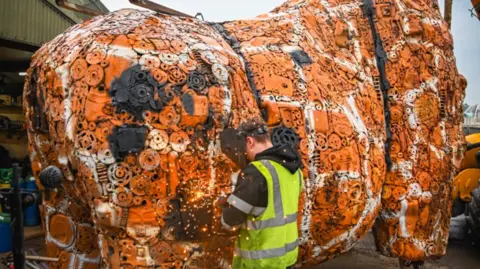 British Ironworks Centre A man with a yellow reflective jacket and face mask facing a large orange and white sculpture, representing the main body of the giraffe, which is made from thousands of car parts.