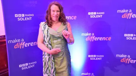 Portia Powell, in a green dress, holding her award, with a purple wall behind her reading "BBC Radio Solent make a difference"