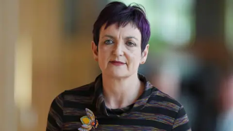 Angela Constance who has short purple hair, wearing a dark stripy top in the Scottish Parliament