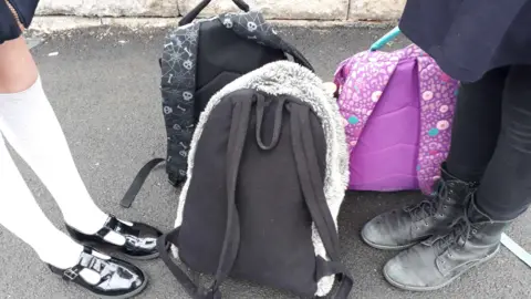 BBC Two sets of feet - one dressed in white socks and black patent shoes, the other in black leggings and black lace-up boots, are standing on the concrete. Three rucksacks, in various colours, are on the floor between their feet. 