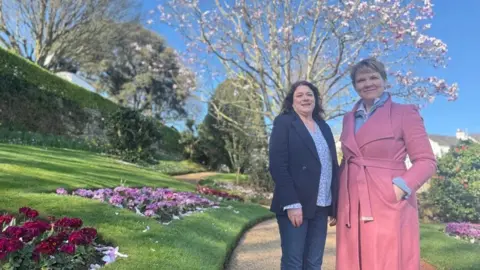 Sue Bacon, Chair of the Floral Guernsey Foundation Council and Deputy Sasha Kazantseva-Miller, President of the Committee for Economic Development are standing in a park. A pink magnolia tree is in bloom behind them. There are also flower beds in a green lawn with dark red and pink flowers in bloom. 