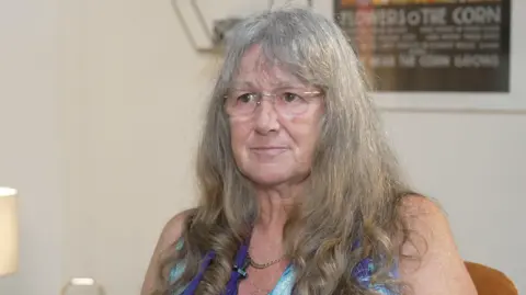 A woman with long greying hair looks seriously off towards the side of the camera. She's wearing frameless glasses and a blue flowered patterned scarf. 