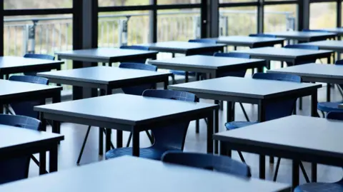 Rows of empty desks with chairs tucked underneath in a room. To the right is a large glass window, with a railing outside. 