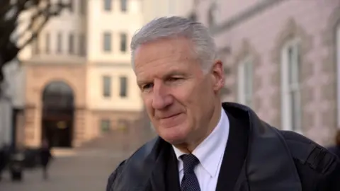 BBC Deputy Binet is wearing a black jacket, a white shirt and a black tie with white polka dots. He has white hair. 