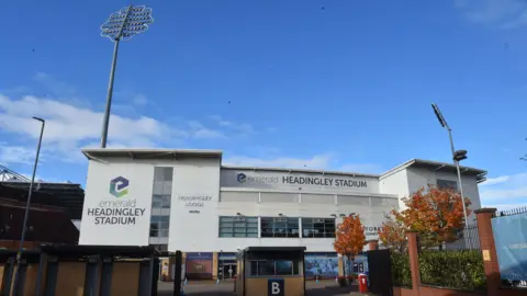 PA Media The exterior of Headingley Stadium is shown with its large white panels, glass windows and tall floodlights rising above the building. A gated entrance and autumn‑coloured trees sit in the foreground, making the stadium look both modern and familiar as a major Leeds sports venue.