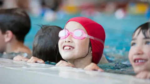A young girl wearing a red cap and pink goggles in the water at the edge of a swimming pool during a swimming lesson. There are three children next to her.