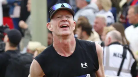 A man is running in a black and orange tank top, with a black hat and blue sunglasses on his head. He has an exhausted expression and in the background there is a large crowd of people behind a barricade.
