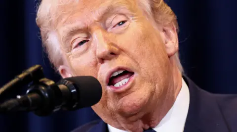 US President Donald Trump speaks into a microphone, his mouth is agape and he is wearing a blue suit jacket and white shirt. Behind him is a dark blue curtain.