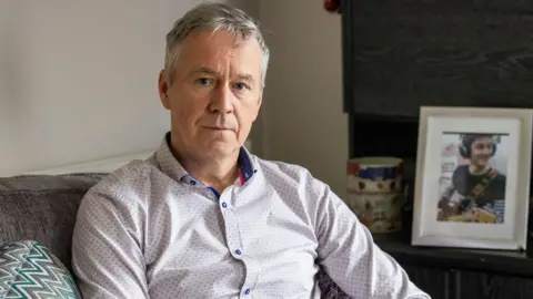 Peter Dolan in a white shirt with a photo of his son behind him. He is sitting on a sofa.