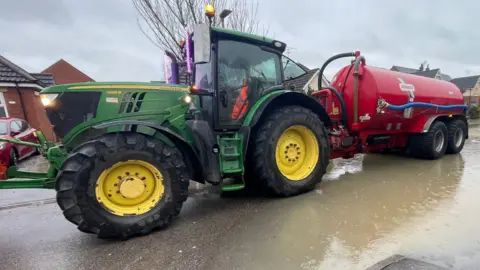 A green tractor with yellow wheels with a black exterior is towing a red tanker. It has its front lights on and flooded water can be seen in a residential street. 