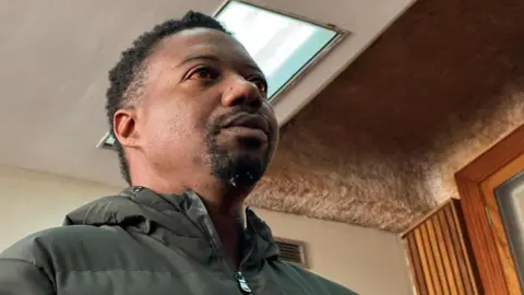 Reuters Ndodana Mkhanyisi Tshuma: The head and shoulders of a man with dark hair and a dark beard. He is wearing a dark green coat with a hood.