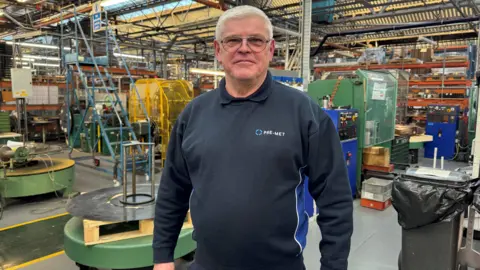 BBC A man wearing a blue jumper, stood on a factory floor - behind him are bits of metal machinery.