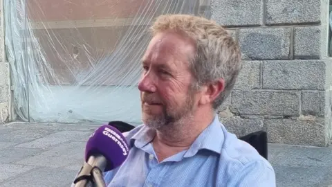 A men sitting behind purple BBC Radio Guernsey microphones. The man is wearing a blue t-shirt with the words inactive on it.