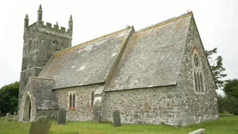 St Grada Church in Cornwall. It is a stone building with a tiled roof in a triangular shape. Several graves are dotted around the outside of the building.