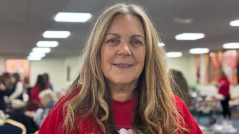 Angela has long honey blonde hair and is wearing a red top. In the background, people are wrapping presents on tables