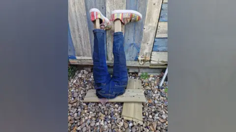 James Coxall A pair of fake legs are placed against a garden shed. They are made from jeans and wooden poles and there are a pair of colourful shoes on the wooden feet.