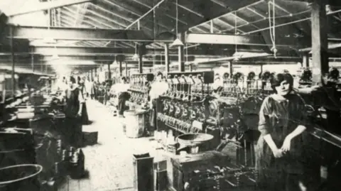 Cheshire Image Bank/Jim and Jennie Lloyd A black and white photograph of inside the BICC factory. A number of workers are standing close cable machine and equipment. 