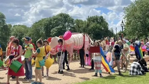 A large group of people dressed in colourful clothing marching through a park in a Pride parade. Some can be seen holding drums, flags and various displays.