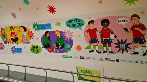 A portion of the mural. It covers part of the enclosed bridge between the interchange and station. Cartoon images of children from different backgrounds are depicted playing with speech bubbles of northern slang - such as "nah then" and "mucka". They are accompanied by information about local support services and QR codes.