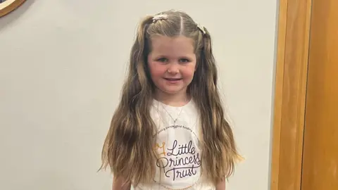 Heidi has her blonde in pigtails that reach down to her elbows. She is wearing a white T-shirt that reads Little Princess Trust.
