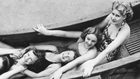 A black and white photo of four women wearing swimwear, lying down in a row on a wooden helterskelter ride and smiling at the camera.