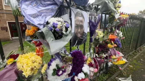 BBC A photo of Smith appears on a fence surrounded by floral tributes outside a house.