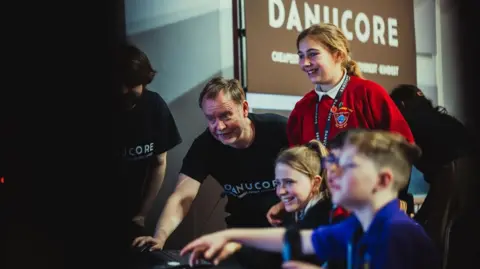 Children crowd around a screen, as two adults demonstrate something to them, they are all smiling and engaged.