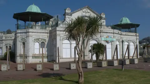 The exterior of Torquay Pavilion which is a white building in a Victorian style. There are two trees in front of it.