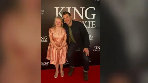 Family Handout Claudia standing next to Peter Coonan. Claudia has long blonde hair and is wearing a pink dress with a pair of gold pumps. Peter is wearing a black jacket, green top, denim jeans and brown boots.