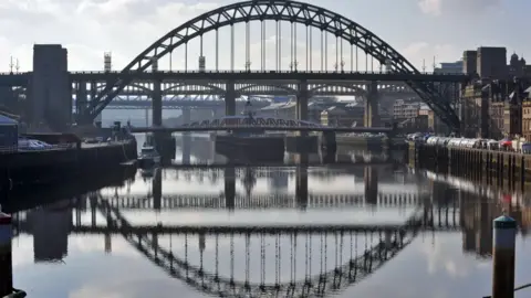 The Tyne Bridge from about 100 metres away looking directly down the river. The bridge's road and steel arch are reflected in the River Tyne. Further bridges can be seen in the distance.