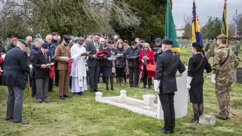 Friends of Nottingham Road Cemetery Three members of uniformed military stand in a graveyard holding flags with their backs to the camera. In front of them is a white headstone. In front of that is a crowd of around fifty people, the majority of them holding an order of service. In the background are many trees.