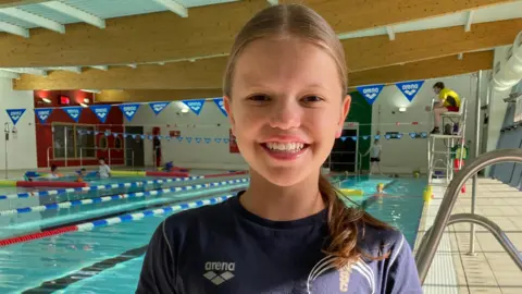 James Burridge/BBC A girl with dark blonde hair in a ponytail pulled over one shoulder, wearing a blue T-shirt and standing in front of a bright blue swimming pool with a life guard on a tall chair to her right and some swimmers to her left