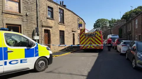 A police van is parked in front of a police cordon - marked with blue and white police tape. A red and yellow fire engine in parked within the cordon in a narrow street with terrace houses.