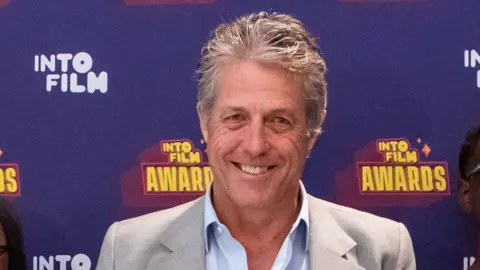 Getty Images Hugh Grant wearing a light grey suit jacket and a light blue top. He has short light greying hair. He is standing in front of a screen that is purple and says INTO FILM AWARDS in yellow.