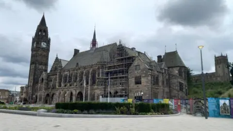 Victorian Gothic building of Rochdale Town Hall with a clock tower to the left and a church on a slope behind it. Part of the town hall is covered in scaffolding for refurbishments.