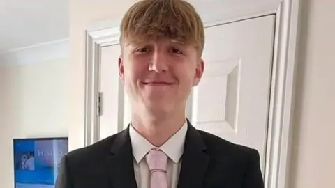 Family handout Alfie Gray. He has short blonde hair and is wearing a white shirt, pink tie and a black suit jacket. He is pictured inside a house in front of a door, looking directly at the camera and smiling.