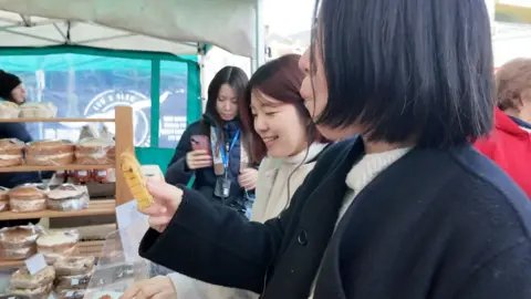 Three women stand in front of a stall at a framers market. They are eating saffron bread that has been cut into slices and handed out. 