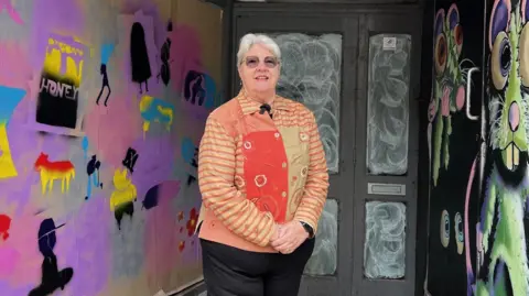 A lady wearing a red and orange patterned jacket stands in front of a doorway covered in colourful street art. The left wall is filled with bright stencil graffiti, while the right wall features a large green cartoon rat possibly thinking about a piece of cheese. The door behind them is frosted with a white pattern.