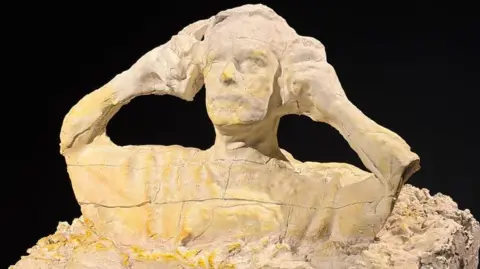 A sculpture of a man putting on ear defenders is made out of plaster. 