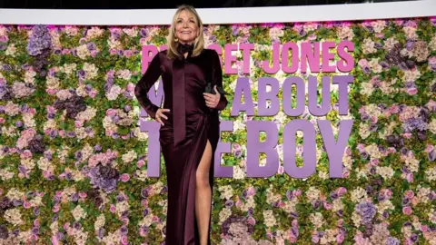 Getty Images Helen Fielding wearing a long burgundy dress with a long slit up the leg, standing in front of a flower wall