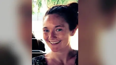 PA Danielle McLaughlin smiling at the camera.  She has long auburn hair tied up in a bun.  She is wearing a dark, patterned top 