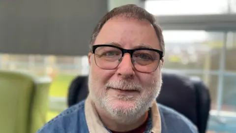 Danny, a man in his 50s, with glasses and a grey beard, wearing a denim shirt